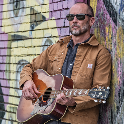 Paul Thorn standing against wall with guitart 